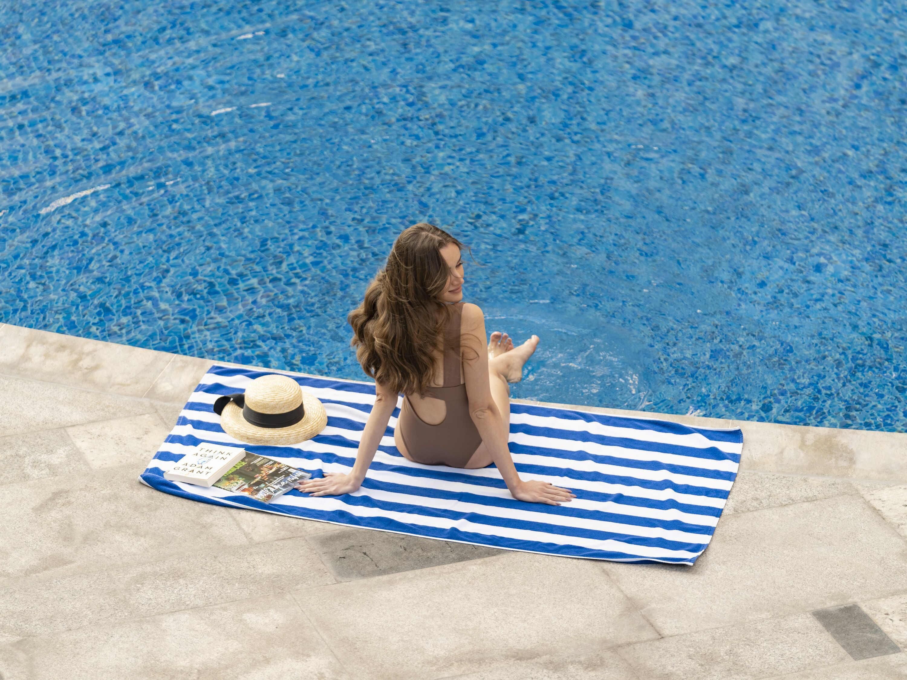 Una persona sentada sobre una toalla azul y blanca a rayas junto a una piscina.
