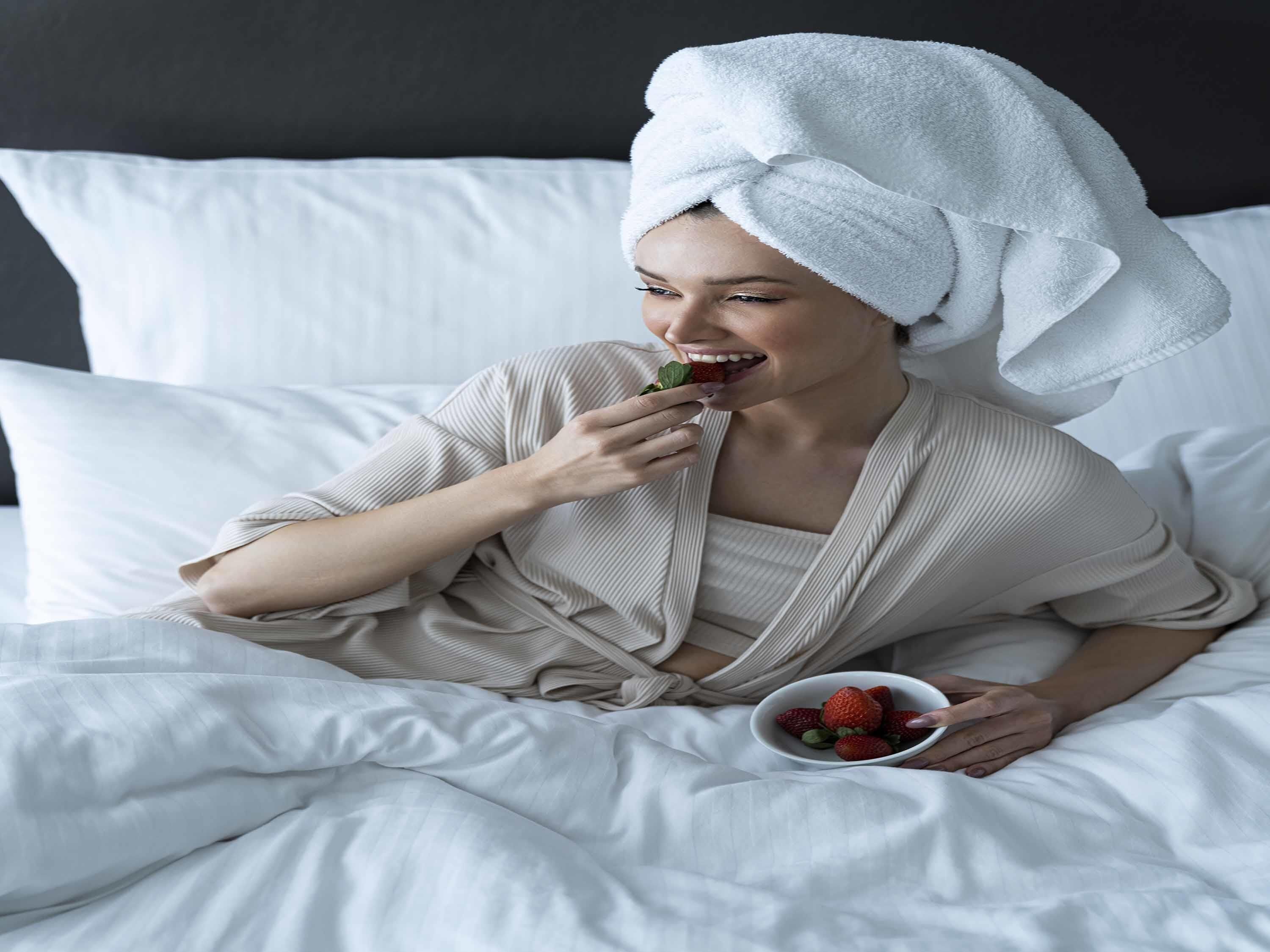 A person sitting on a bed with white bedding, wearing a beige robe, and holding a bowl of strawberries.