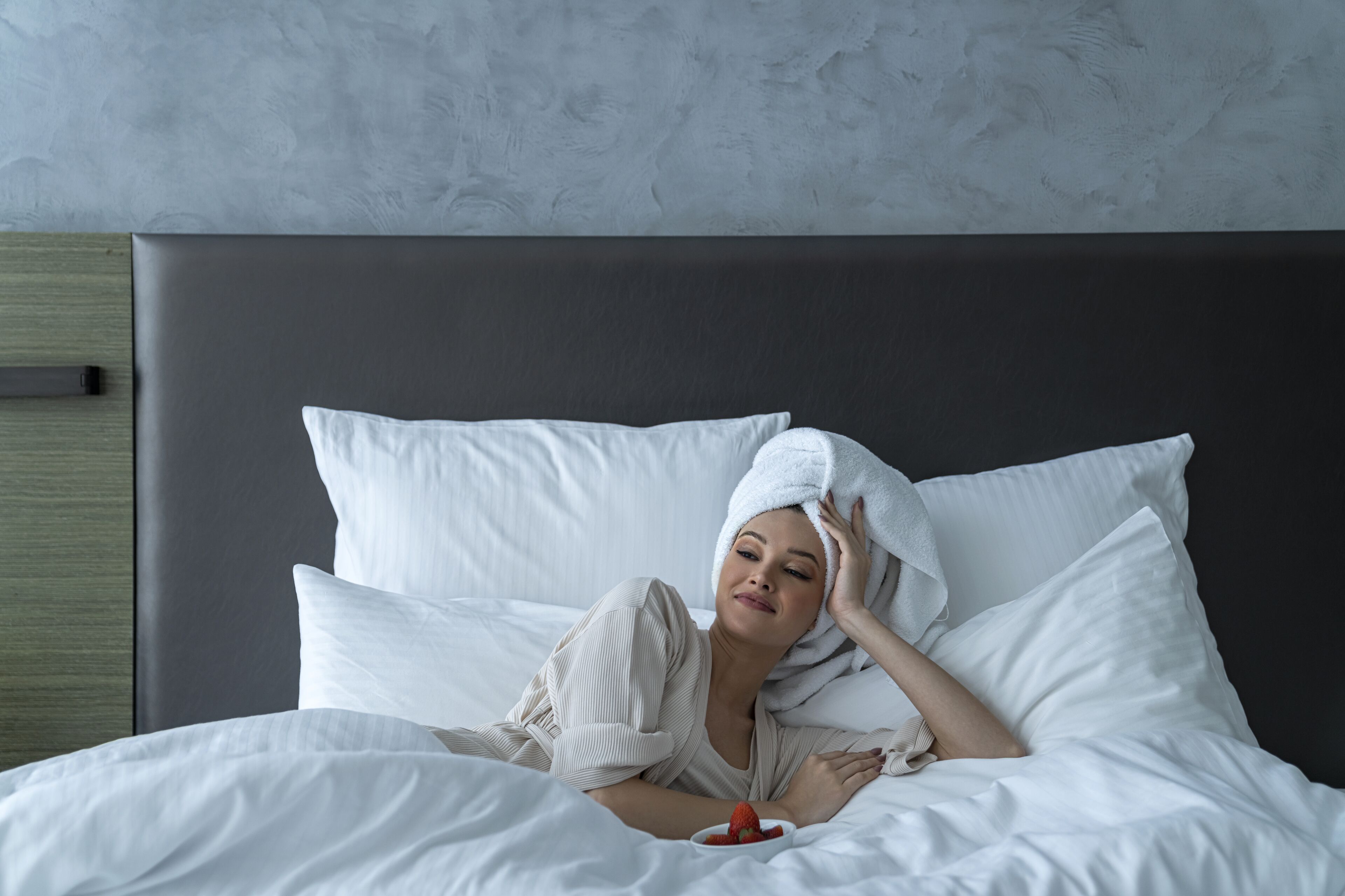 Uma pessoa deitada em uma cama com roupa de cama e travesseiros brancos, uma tigela de morangos e uma cabeceira cinza.