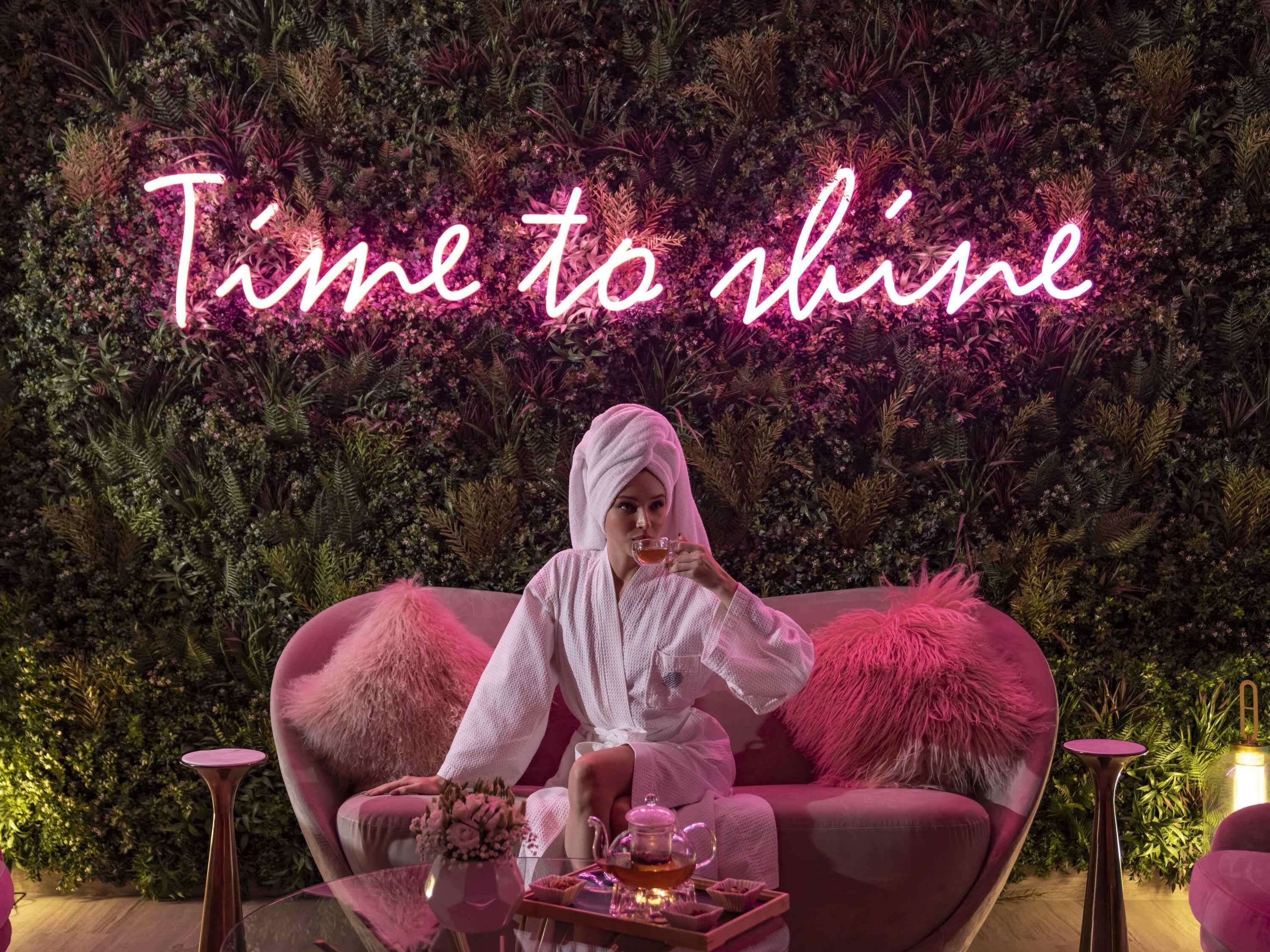 Uma pessoa sentada num sofá rosa, com um letreiro neon que diz 'Time to shine' ao fundo.
