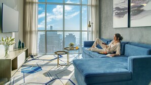 A modern living room with a large window, a blue sofa, a small side table, and a vase with flowers. - Pullman Sharjah (Sharjah)