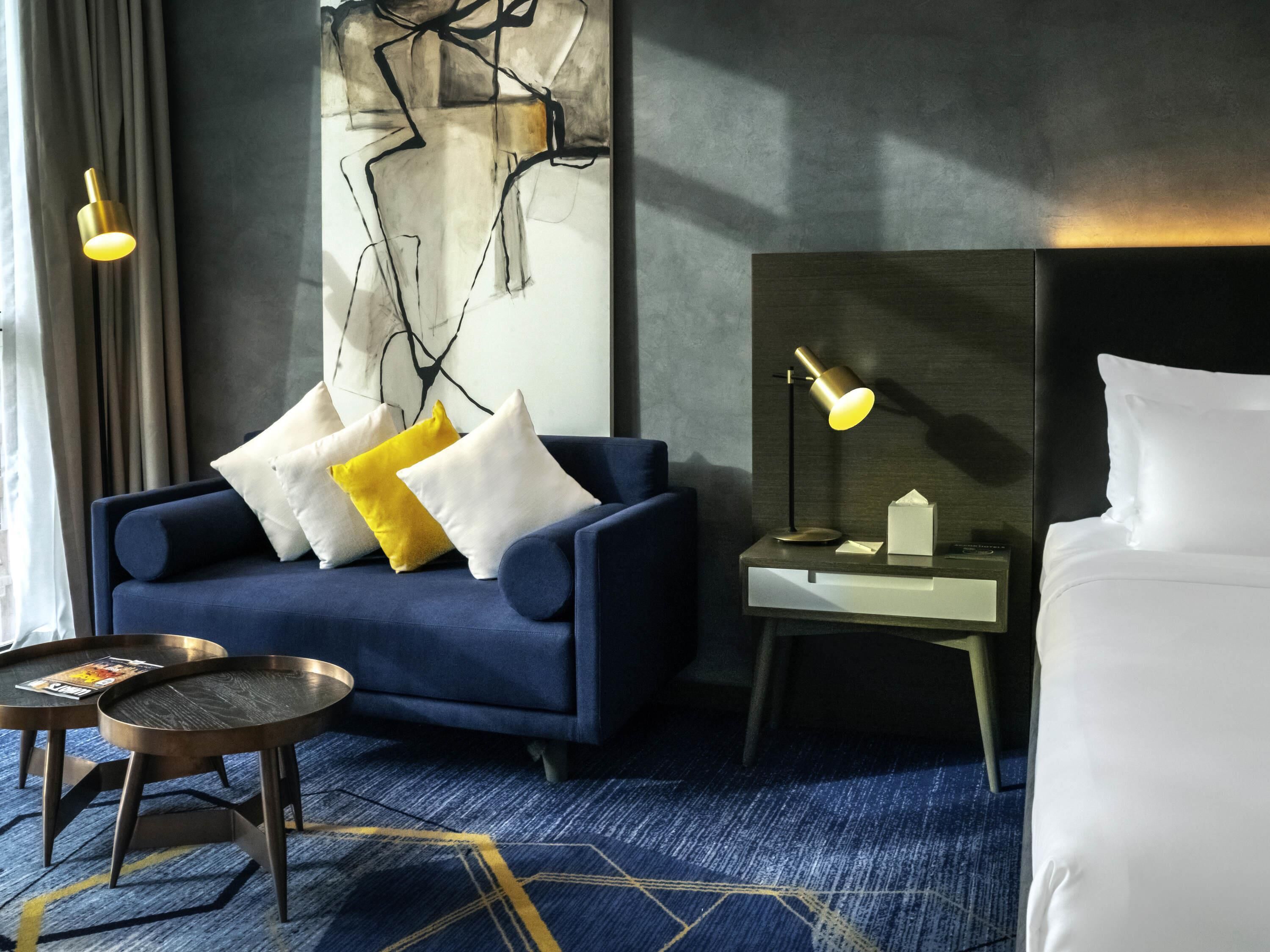 A modern hotel room with a blue sofa, a bedside table with a lamp, a bed with white linens, and a geometric-patterned carpet.