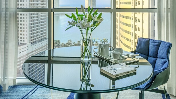 A modern room with a glass table, a vase with flowers, a chair, and a view of tall buildings and a body of water. - Pullman Sharjah (Sharjah)