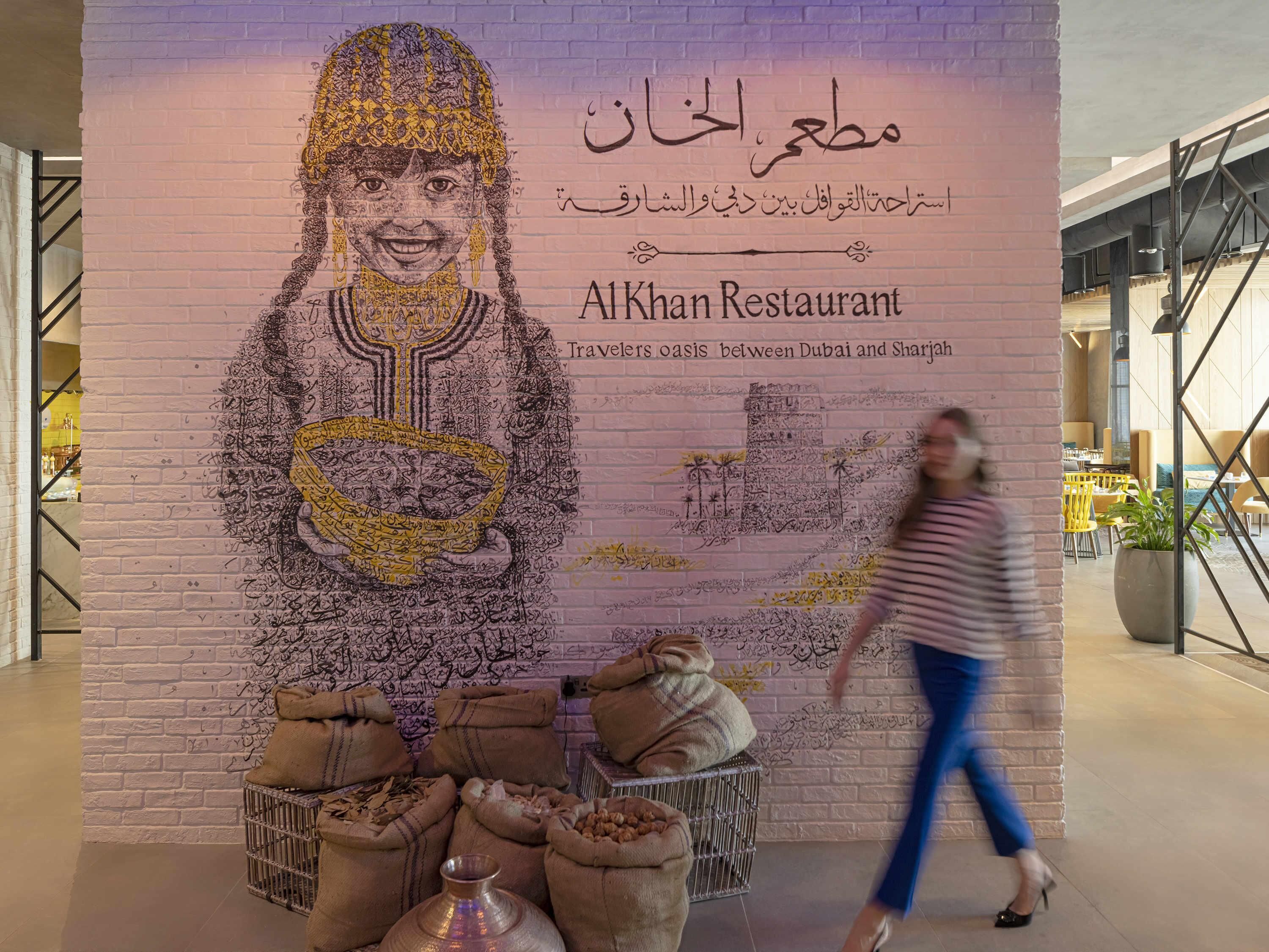 Un mur orné d’une fresque représentant une personne tenant un bol, avec du texte en arabe et le nom « Al Khan Restaurant », ainsi qu’un slogan.