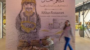 Un mur orné d’une fresque représentant une personne tenant un bol, avec du texte en arabe et le nom « Al Khan Restaurant », ainsi qu’un slogan.