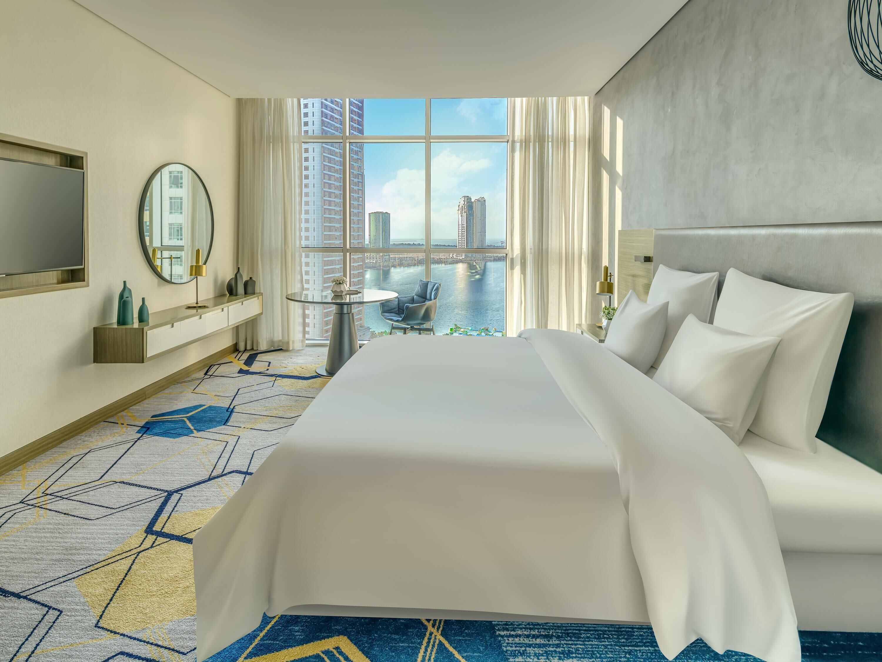 A hotel room with a large bed, a round mirror, a small table, a TV, and a view of a cityscape and water.