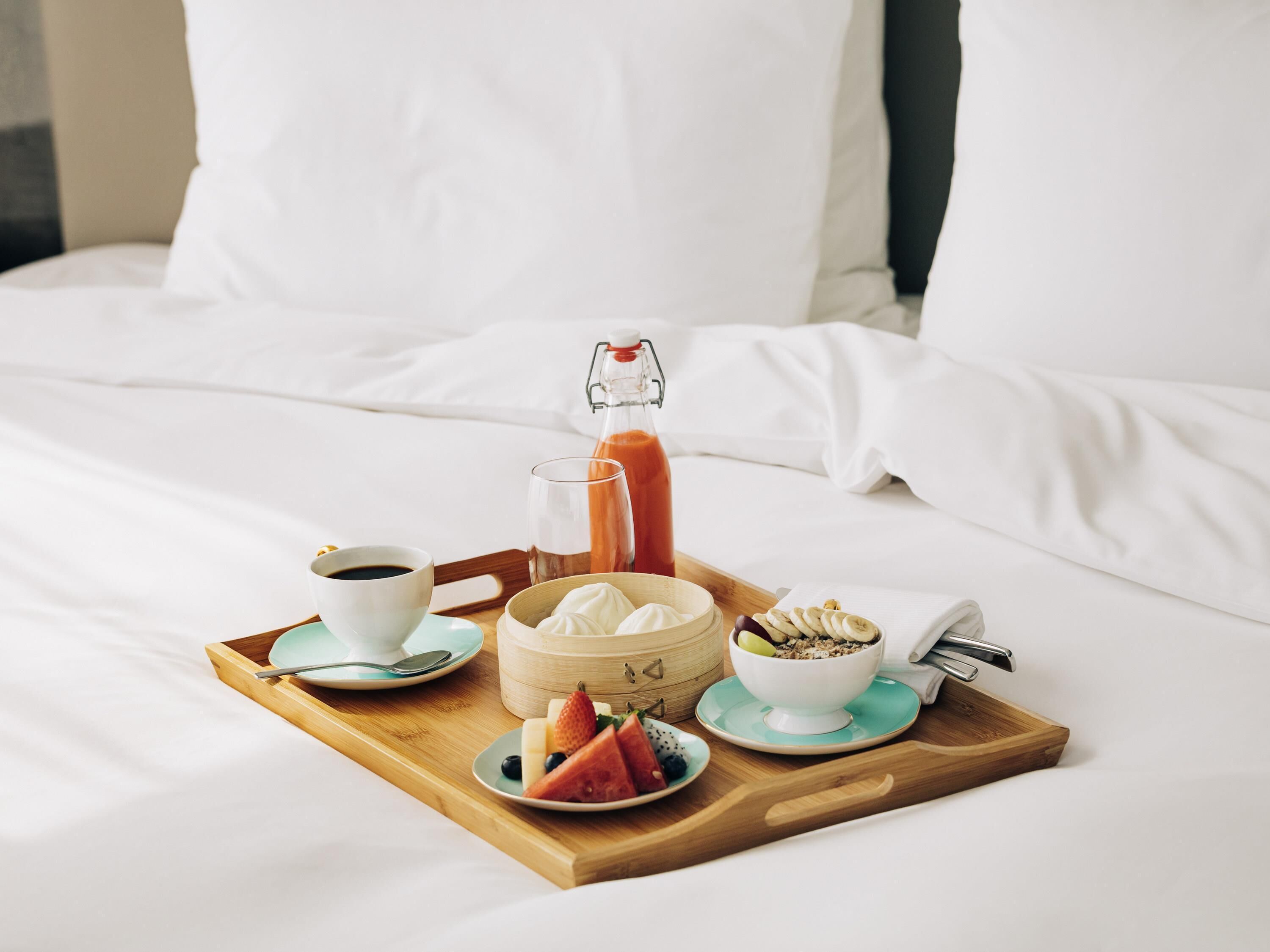 Bandeja de desayuno sobre una cama, con una botella de jugo de naranja, un tazón de yogur, un plato de fruta y una taza de café.