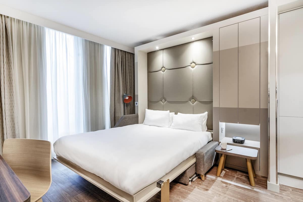 A modern hotel room with a large bed, a bedside table, a chair, and a wardrobe.