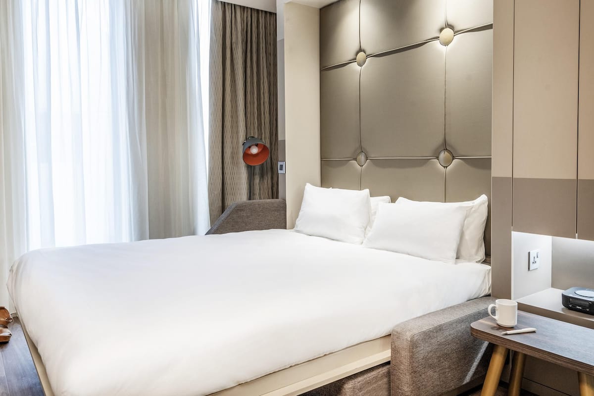 A hotel room with a large bed, a bedside table with a cup and a remote, and a wall-mounted lamp.