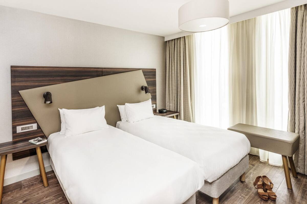 A hotel room with two beds, a large window with curtains, a bedside table with a lamp, and a bench.