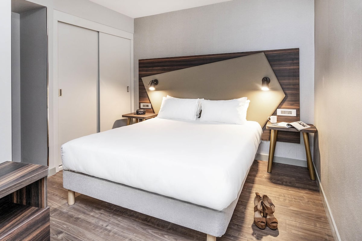 A modern hotel room with a large bed, wooden headboard, bedside tables, and a pair of shoes on the floor.