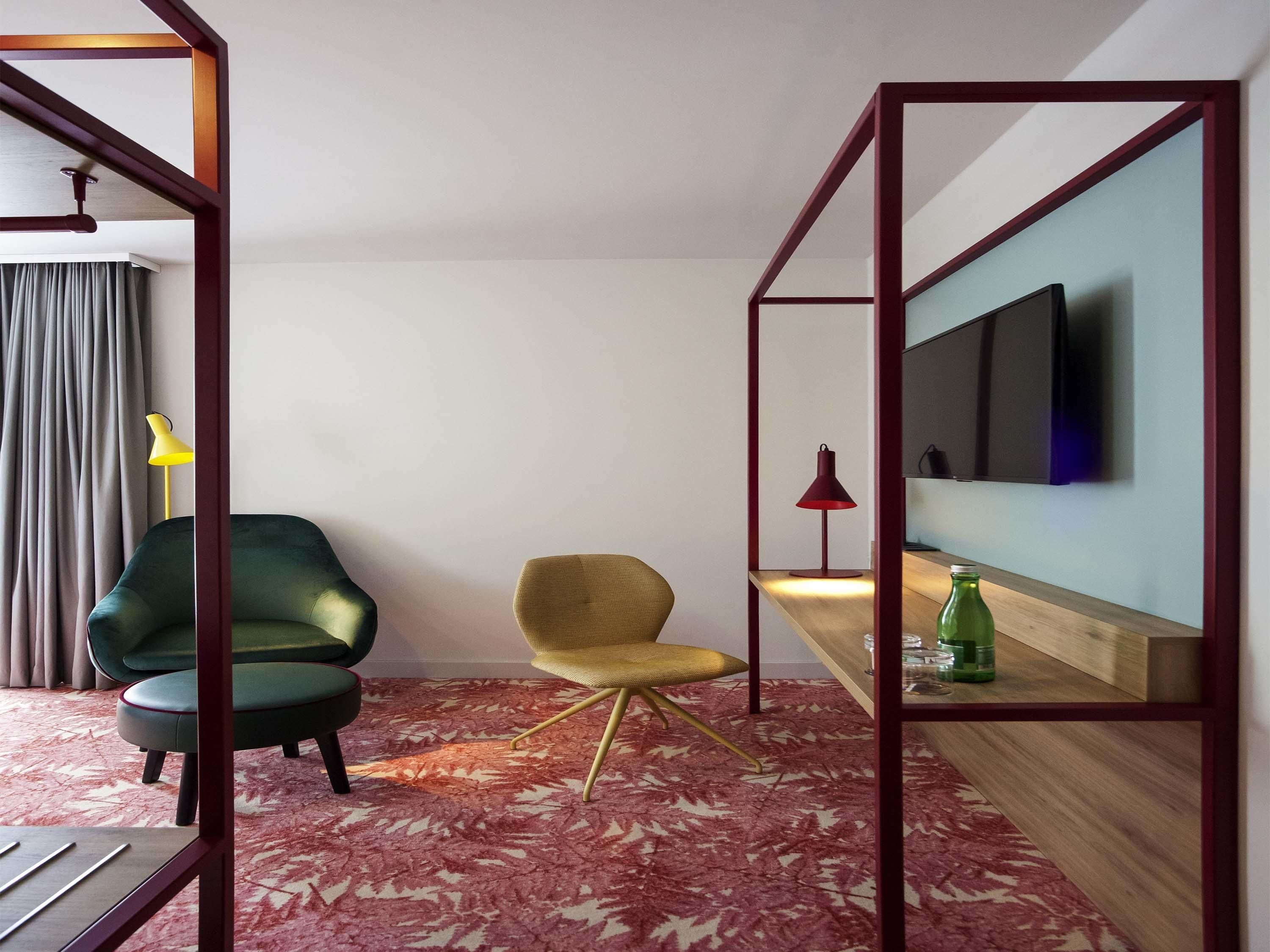 A hotel room with a modern design, featuring a green armchair, a yellow lamp, a red floor lamp, a wooden side table with a bottle, and a TV mounted on the wall.