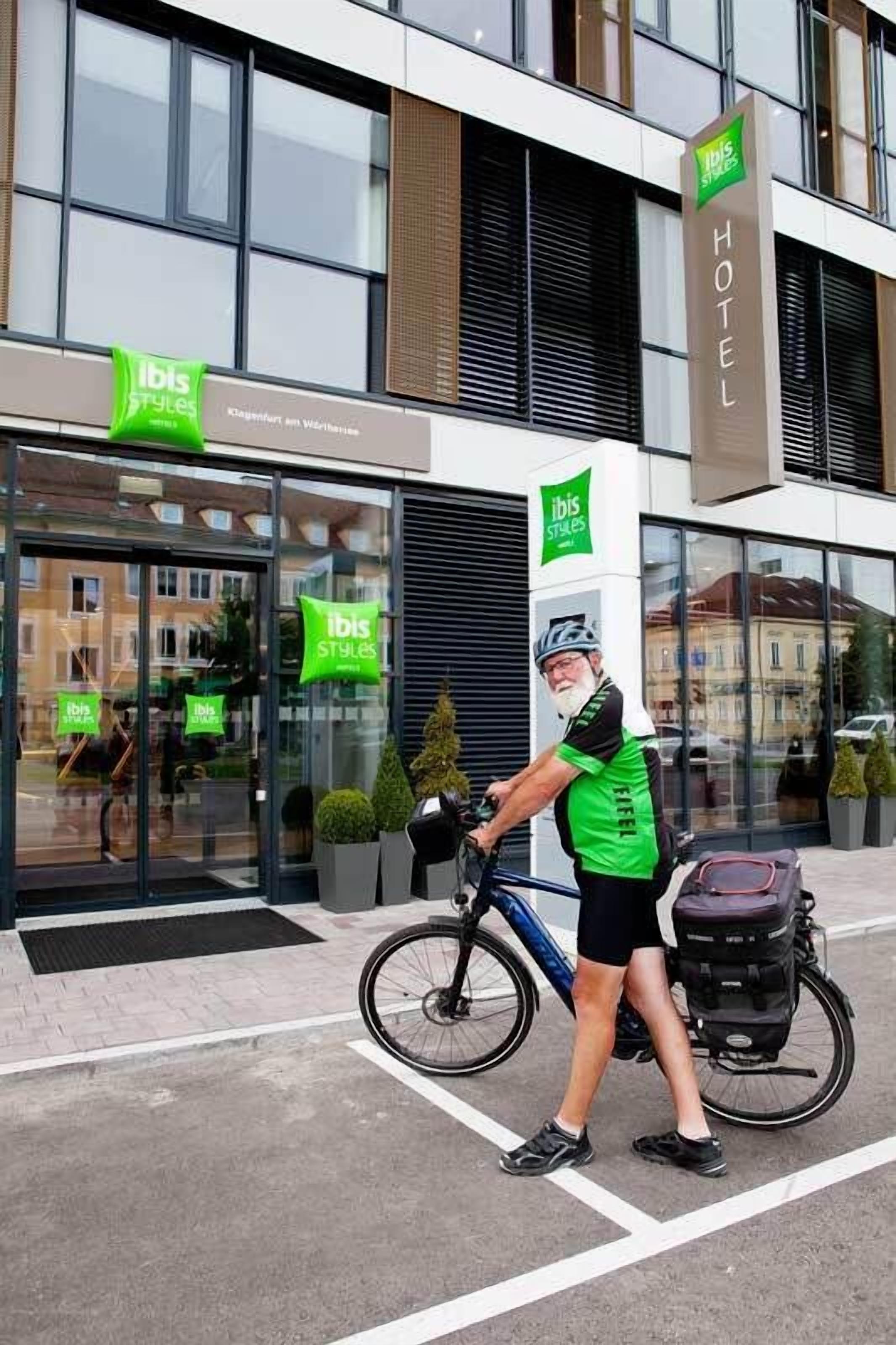 En hotellentrél med en glasfasad, en skylt med texten ”ibis Styles”, och en person med en cykel och en ryggsäck.