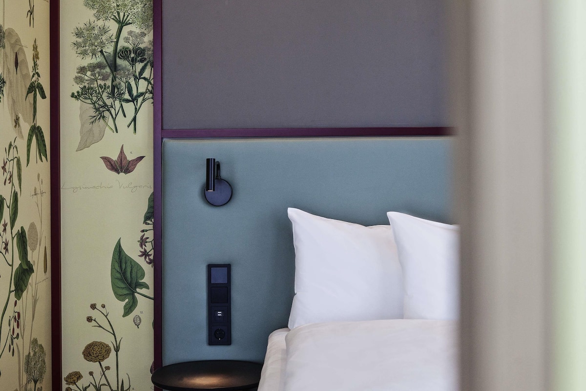 Une chambre à coucher avec un lit, des oreillers blancs, une table de chevet, une lampe et un interrupteur fixé au mur.