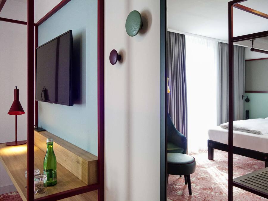 Une chambre d’hôtel avec un lit, un téléviseur fixé au mur, une table de chevet avec une bouteille et des verres, et une chaise.