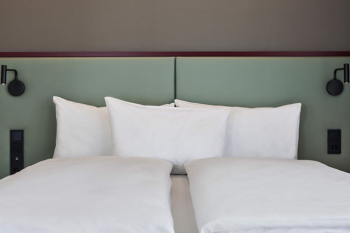 Un lit double avec une literie blanche, une tête de lit verte et deux lampes fixées au mur.