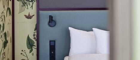 A bedroom with a bed, white pillows, a bedside table, a lamp, and a wall-mounted switch.