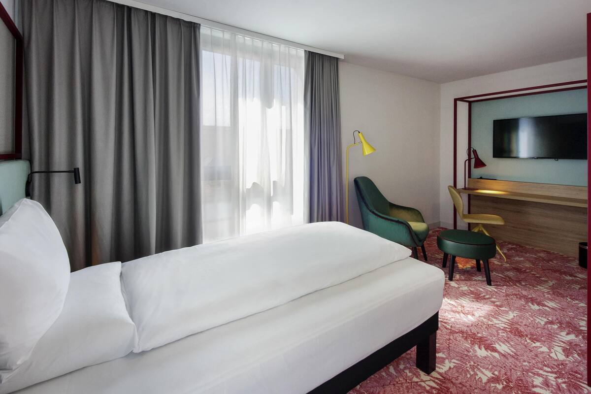 Une chambre d’hôtel avec un lit, un bureau, une chaise, une télévision et une fenêtre avec des rideaux.