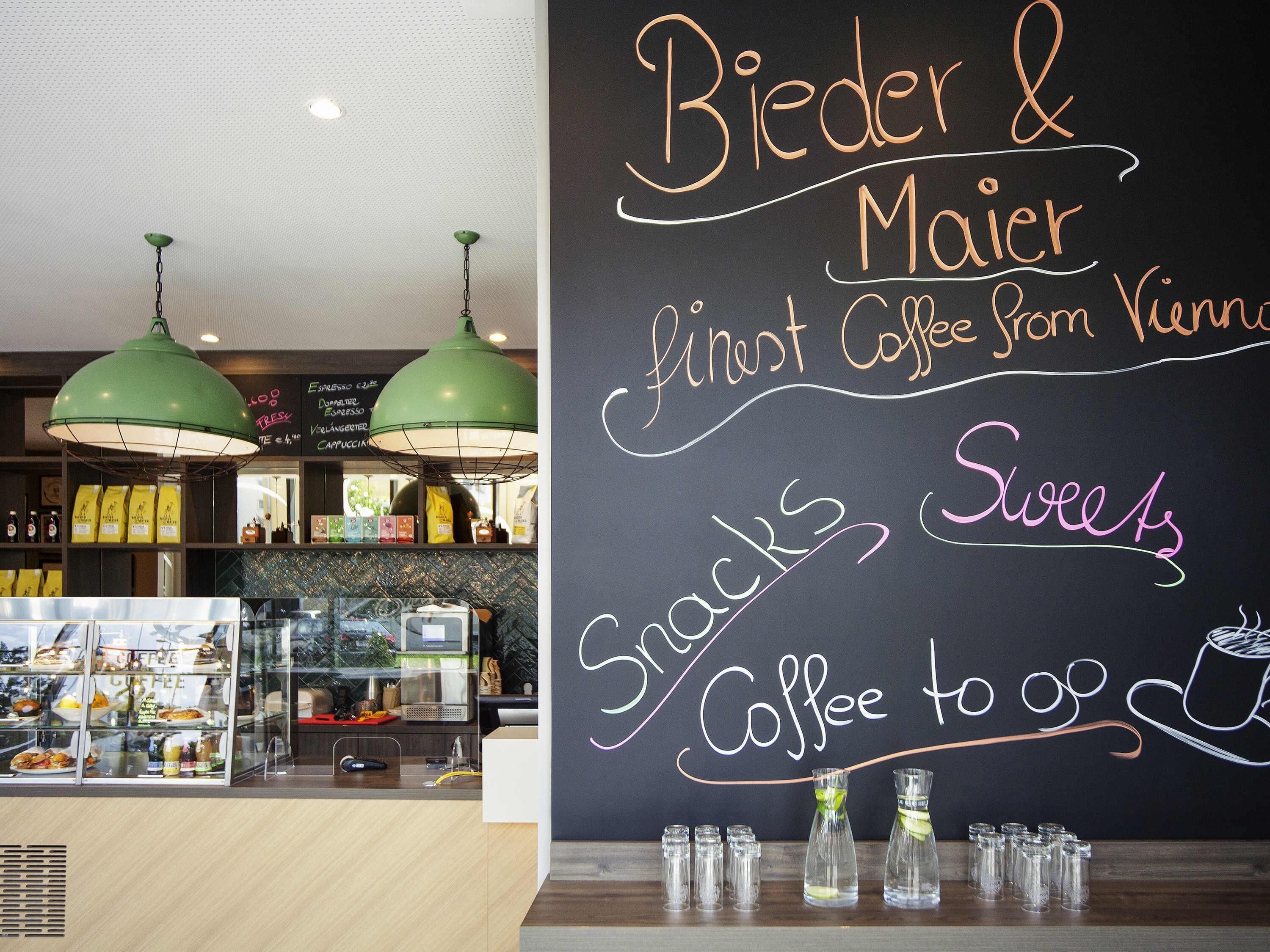 L’intérieur d’un café, avec un tableau noir servant de menu, des suspensions vertes décoratives et un présentoir contenant des collations et des boissons.