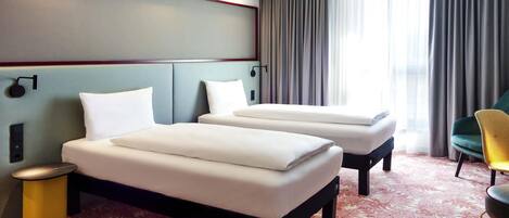 Una habitación de hotel con dos camas, una mesita auxiliar, una silla y un ventanal con cortinas.