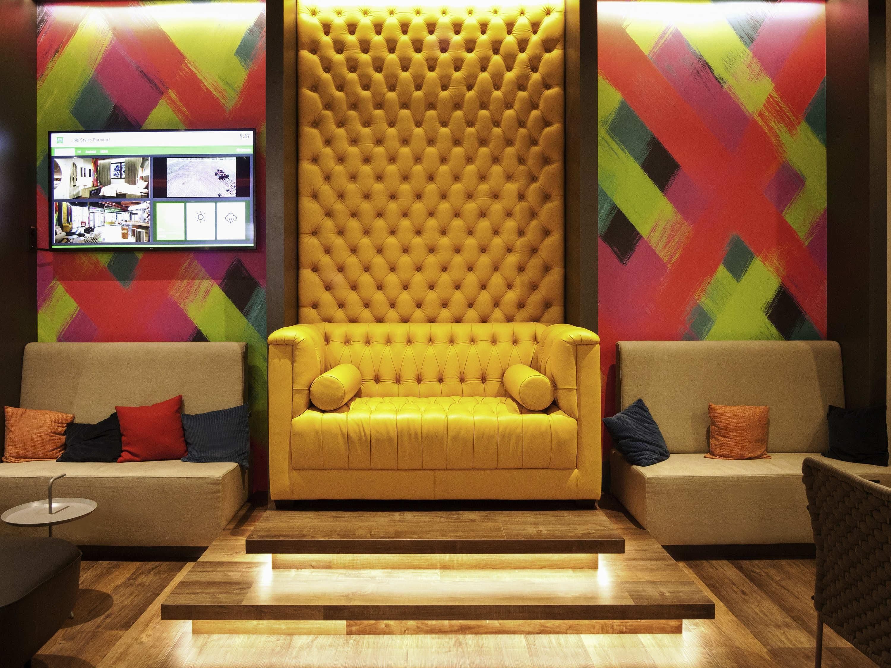 Een moderne lobby met een geel gestofte bank, twee banken met kussens, een houten vloer en kleurrijke geometrische muurkunst.