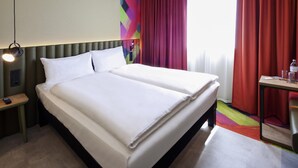 A double bed with white linens, a bedside table with a phone, a lamp, and a colorful wall design. - ibis Styles Parndorf Neusiedler See (Parndorf)