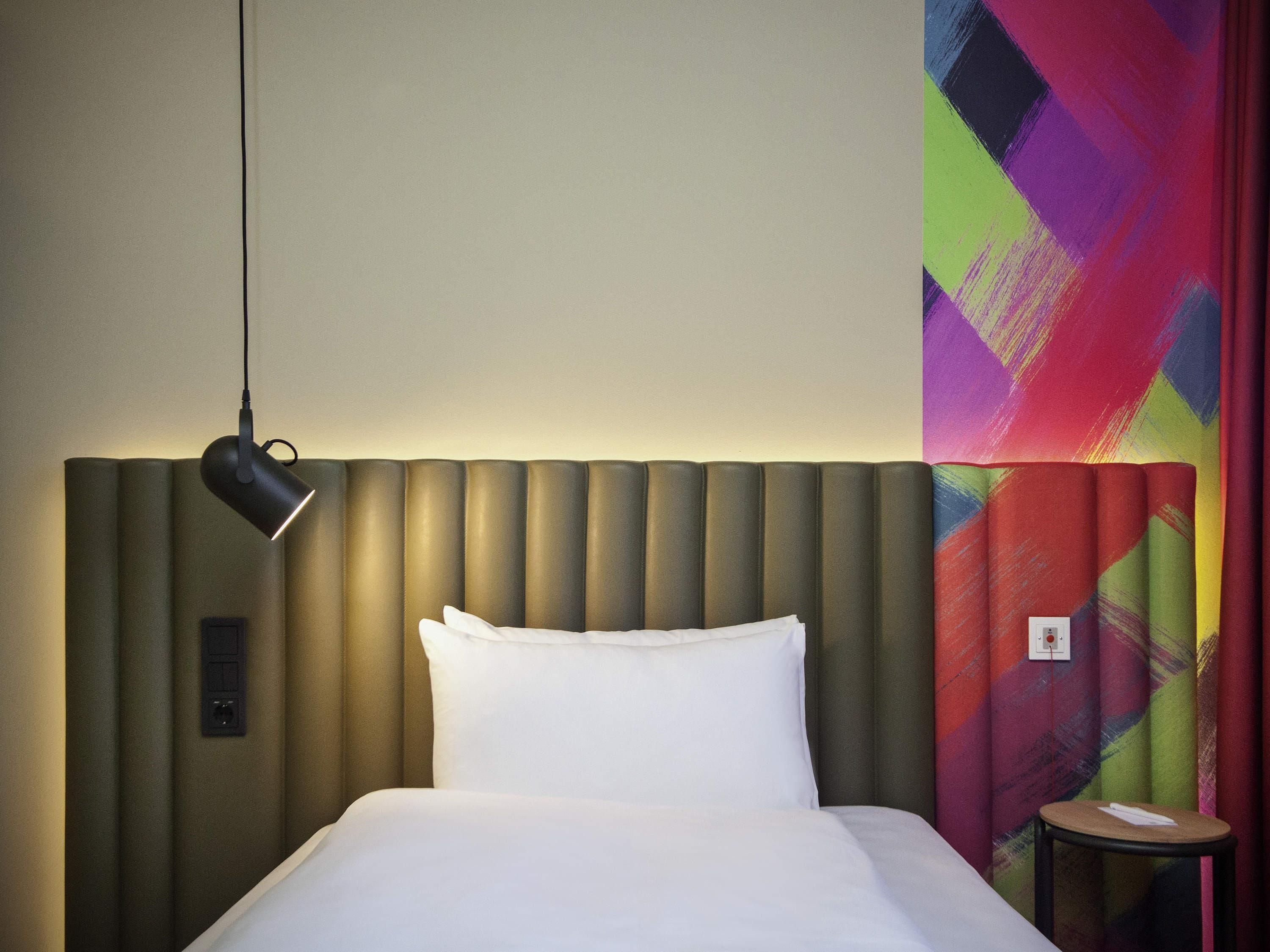 A hotel room with a bed, a bedside lamp, a small table, and a colorful abstract wall.