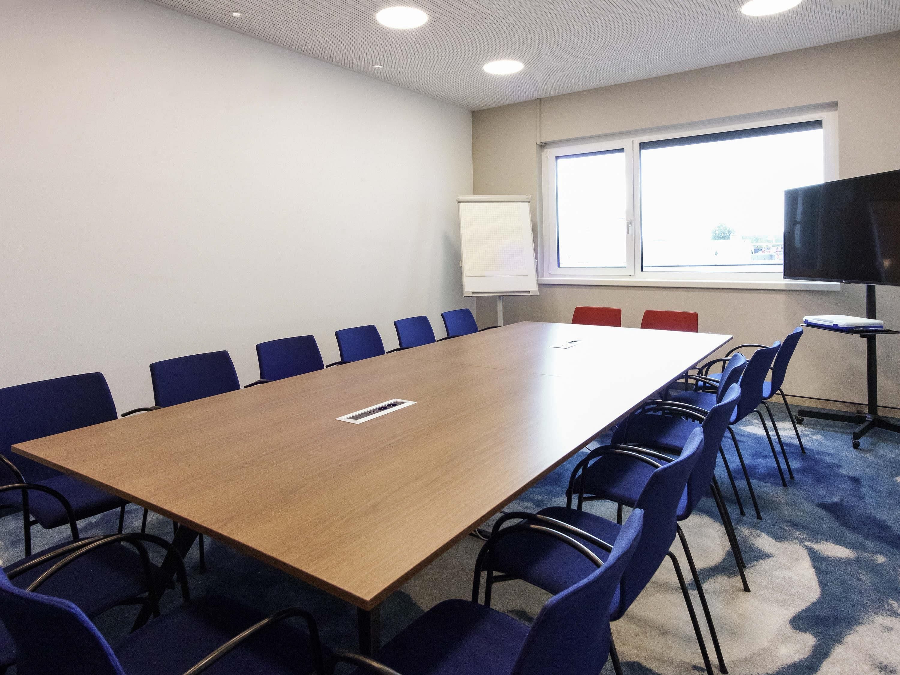 A conference room with a long table, blue chairs, a whiteboard, and a TV on a stand.