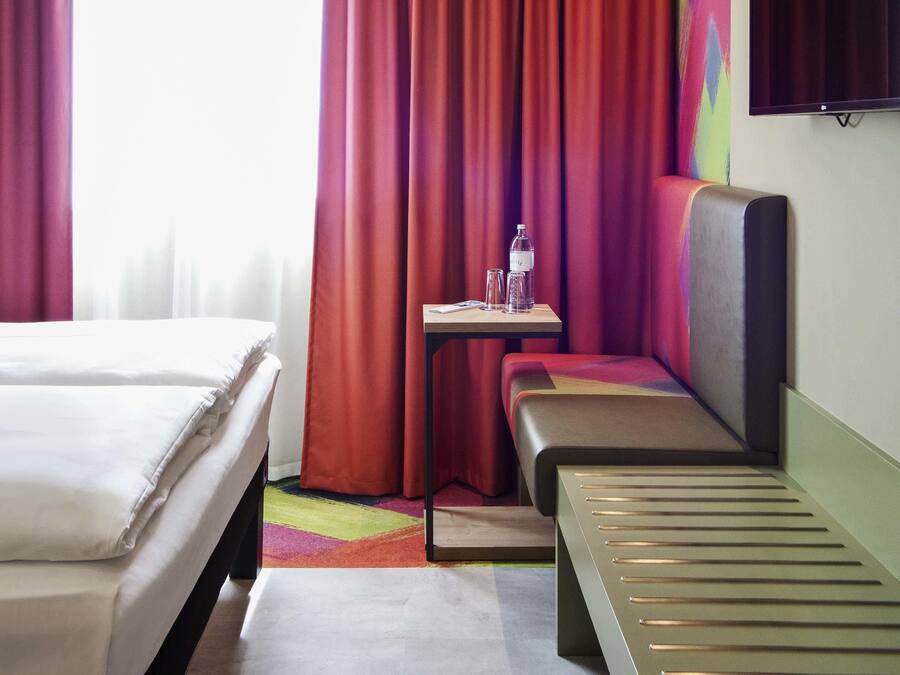 Una habitación de hotel con una cama, un banco, una mesita con una botella y vasos, y un televisor en la pared.