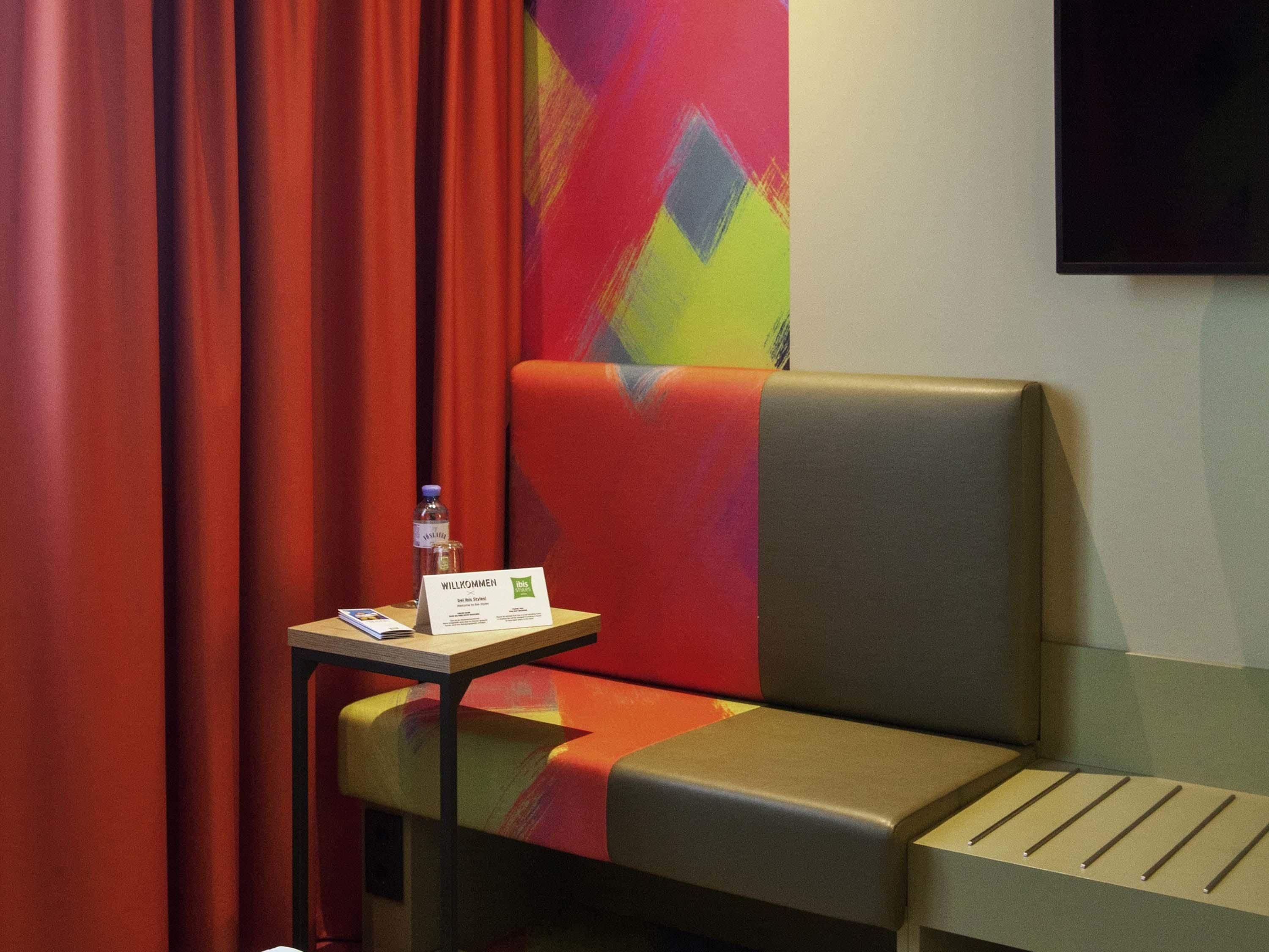 A hotel room with a single sofa, a small table with a welcome sign, a bottle of water, and a colorful wall.