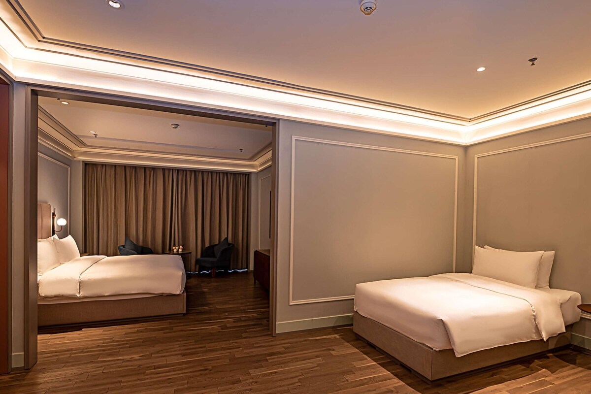 A hotel room with two beds, a sitting area with a chair and a table, and a large window with curtains.
