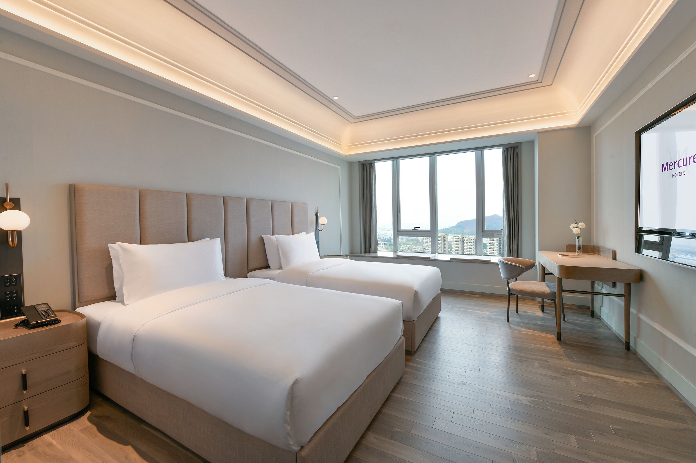 A hotel room with two beds, a desk, a chair, and a large window with a city view.