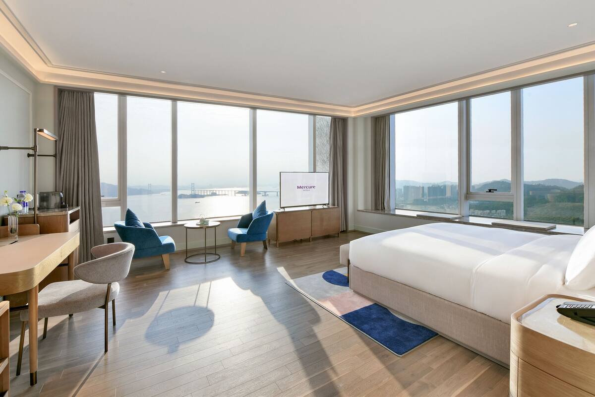 Quarto de hotel moderno com uma cama grande, uma escrivaninha, uma cadeira, uma TV e vista para uma ponte e o horizonte da cidade.