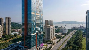 Un hôtel de grande hauteur à la façade moderne en verre, situé près d’une promenade en bord de mer, à proximité d’une autoroute et d’espaces verts.