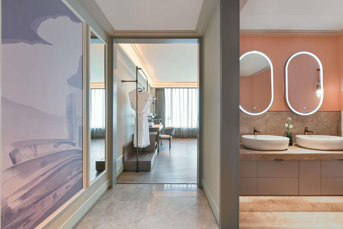 A modern hotel bathroom with two sinks, a large mirror, and a view of a cityscape through a glass door.