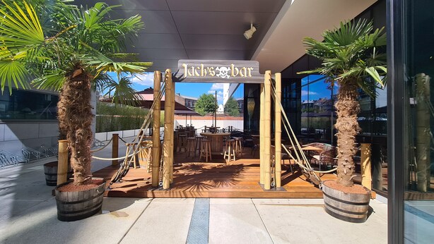 An outdoor seating area with wooden tables and chairs, surrounded by potted plants and bamboo structures. - ibis Styles Liege Guillemins (Liege)