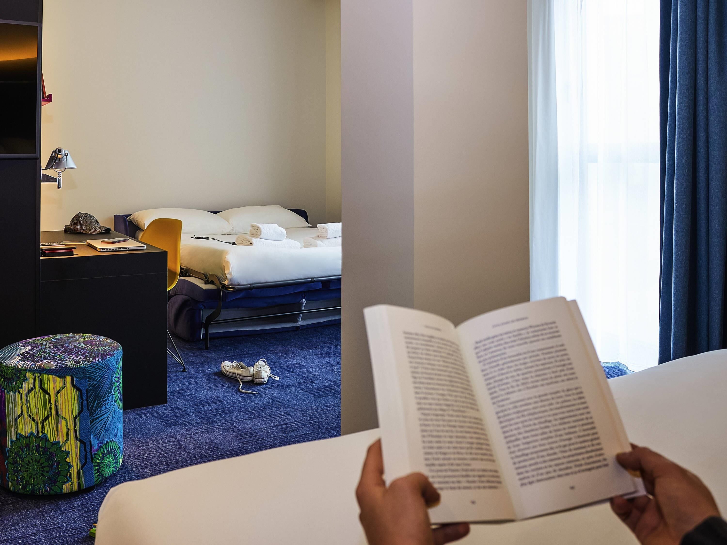 Een hotelkamer met een bed, nachtkastje, lamp en een kleurrijk poef.