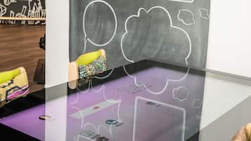 Una habitación con una mesa de vidrio que muestra una interfaz digital, una silla con un cojín de colores y una pared con dibujos abstractos.