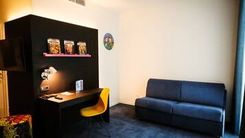 A small, modern room with a dark blue sofa, a desk with a chair, a TV, and a shelf with books.