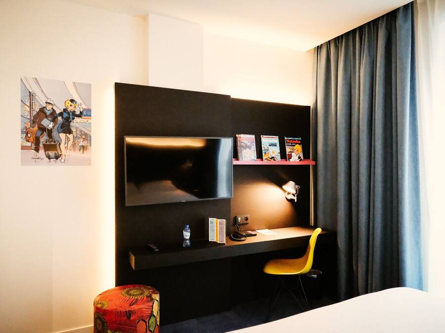Een hotelkamer met een bed, een bureau met stoel, een televisie, een plank met boeken en een raam met gordijnen.