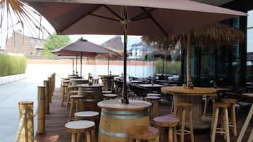An outdoor seating area with wooden tables and stools, umbrellas, and a thatched roof structure.