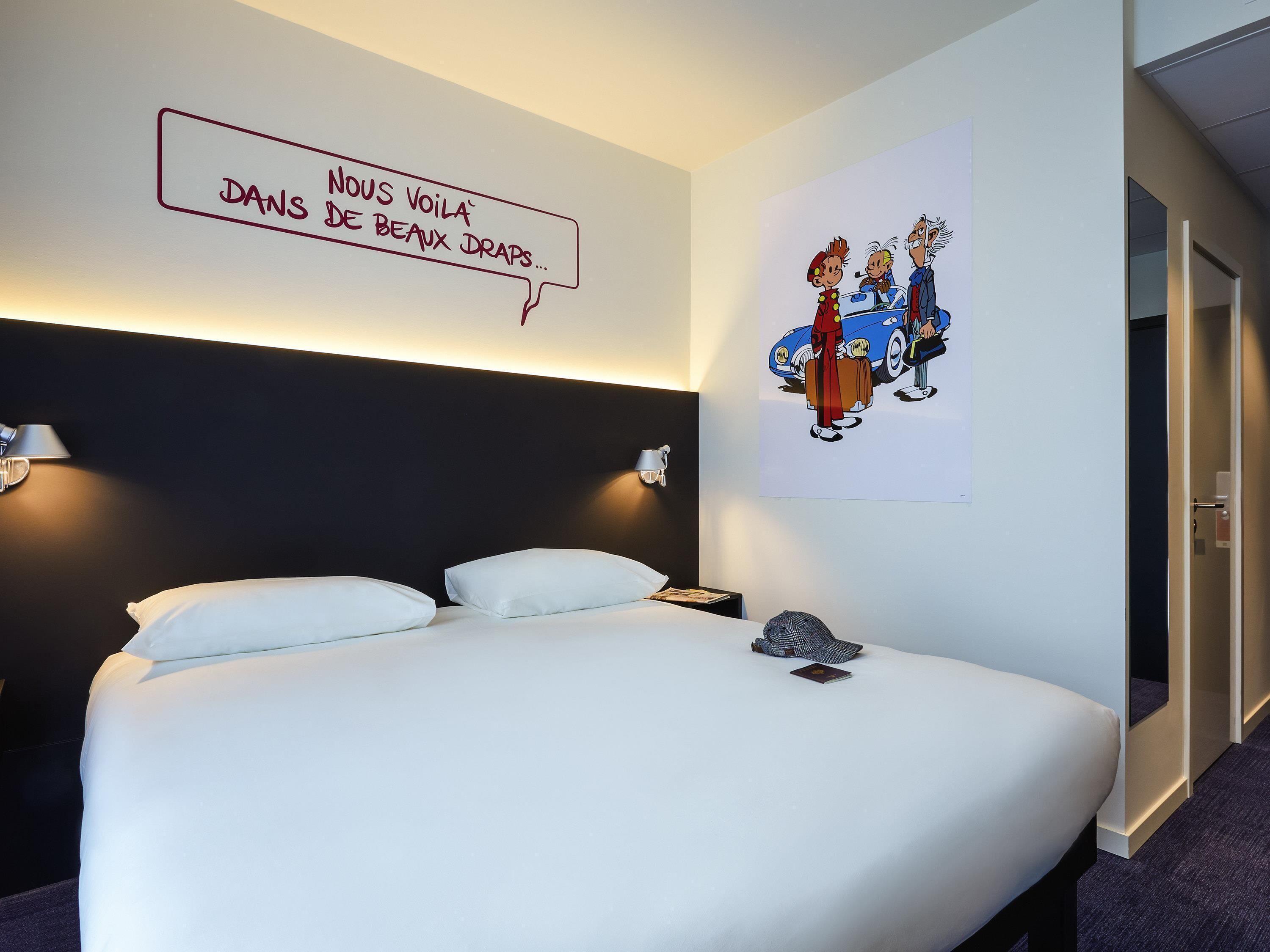 Una habitación de hotel con una cama, dos lámparas de noche, una obra de arte en la pared y una puerta.