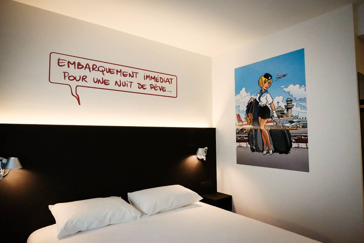 Une chambre d’hôtel avec un lit, un mur orné d’une illustration de style bande dessinée et un panneau avec du texte en français.
