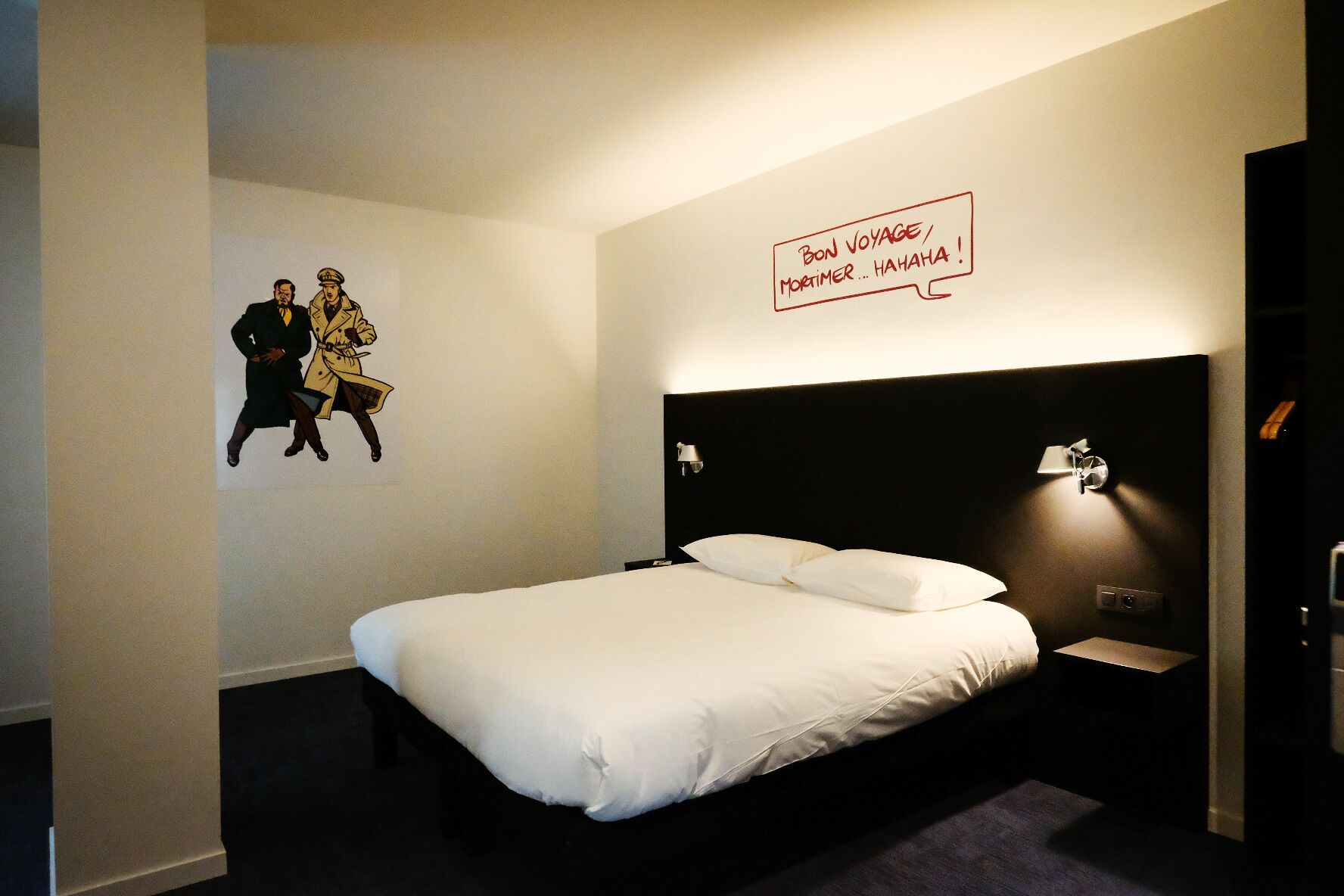 A hotel room with a large bed, a headboard with a light fixture, a wall-mounted artwork, and a small bedside table with a lamp.