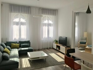 A modern living room with a sofa, coffee table, TV, and dining area. - Paulay Central Top Suite (Budapest)