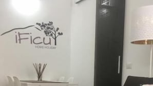 A living room with a brown sofa, a white coffee table, and a dining table with chairs. There is a wall with a logo and text that reads "iFicu HOME HOLIDAY". - iFicu (Catania)