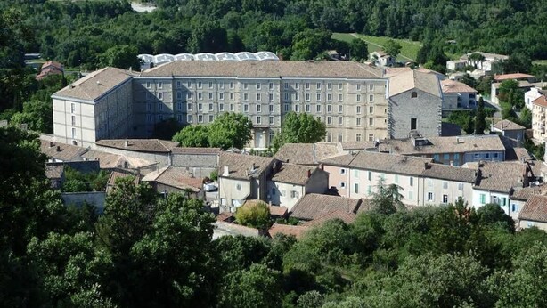 A large, historic building with multiple wings and a central courtyard, surrounded by lush greenery and a river in the background. - Hostellerie Charles de Foucauld (Viviers)