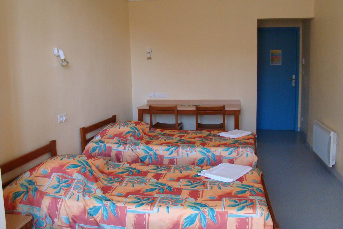 A small, simple room with two single beds, a wooden desk, and a blue door.