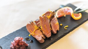 A black slate plate with sliced meat garnished with orange zest, accompanied by a citrus slice and a small piece of red garnish. - Agriturismo Villa Gelso (Vesime)