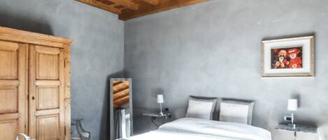 Un dormitorio con techo de madera, una cama con ropa de cama blanca, una mesita de noche con lámpara, un armario y un cuadro en la pared.